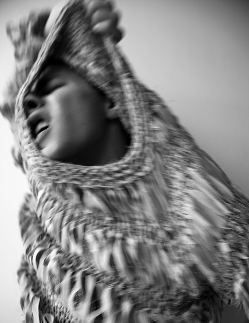 Monochrome portrait of person with sweater partly pulled over their head, eyes closed and mouth open in an indoor setting.
