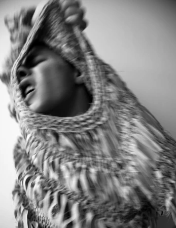Monochrome portrait of person with sweater partly pulled over their head, eyes closed and mouth open in an indoor setting.