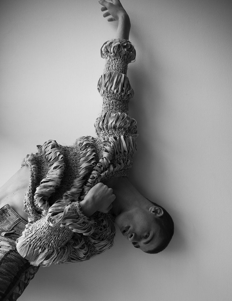 Monochromatic portrait of a person in a sculptural, textured garment, leaning against a wall with one arm raised in a minimalist setting.