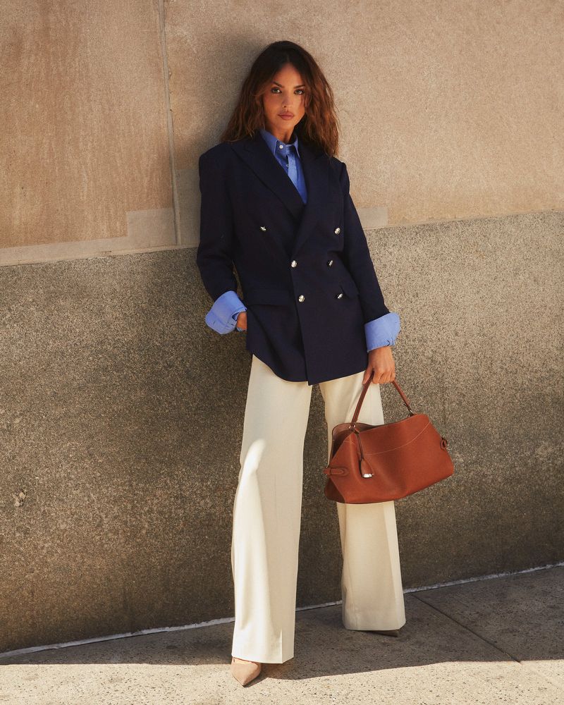 Brunette woman in blazer and wide-leg pants poses against a wall, holding a leather bag. Fashion editorial, outdoor setting.