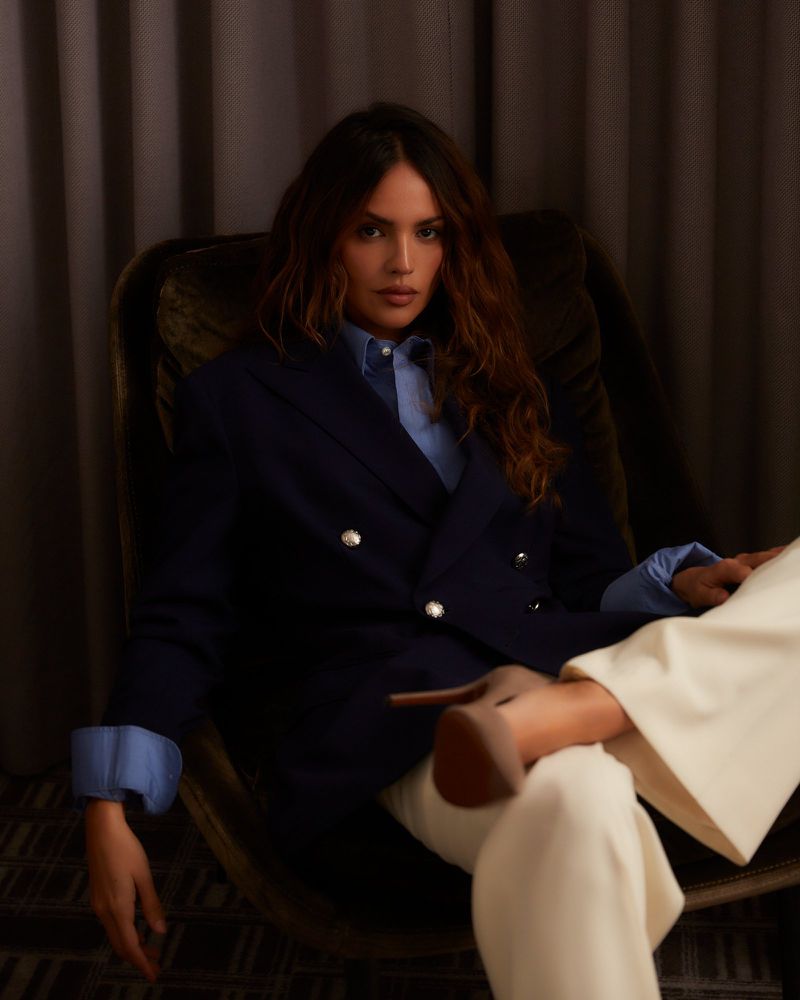 A woman with long brown hair sits in a dark green chair, wearing a blue blazer and cream-colored pants, photographed indoors.