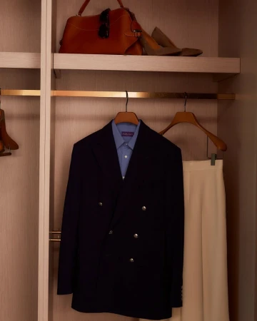 Open closet with navy blue blazer, light blue shirt, and cream-colored trousers hanging on hangers, plus a brown leather handbag on a shelf.