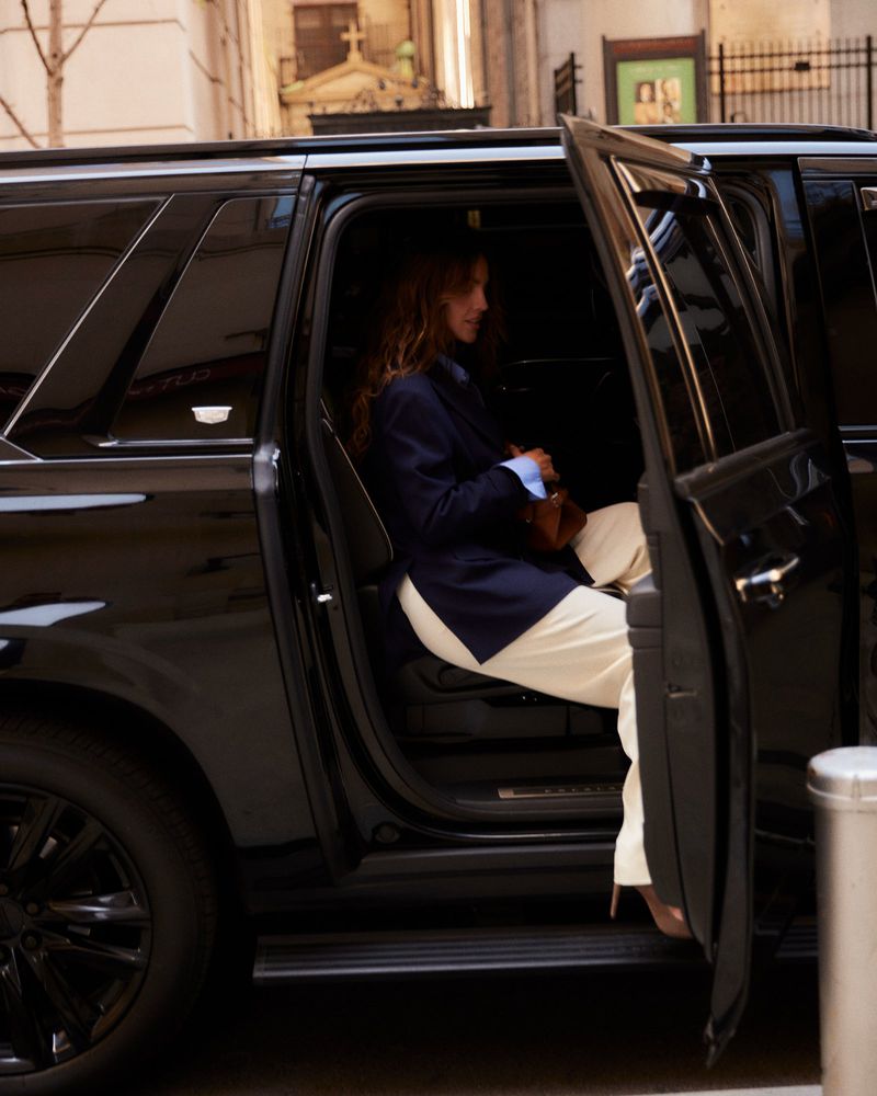 Stylish woman exiting black SUV in city setting, wearing a blazer and light-colored pants. Buildings are visible in the background.