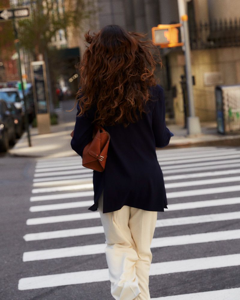Woman with curly hair walks across crosswalk in an urban setting. She carries a brown bag and wears a black jacket and tan pants.