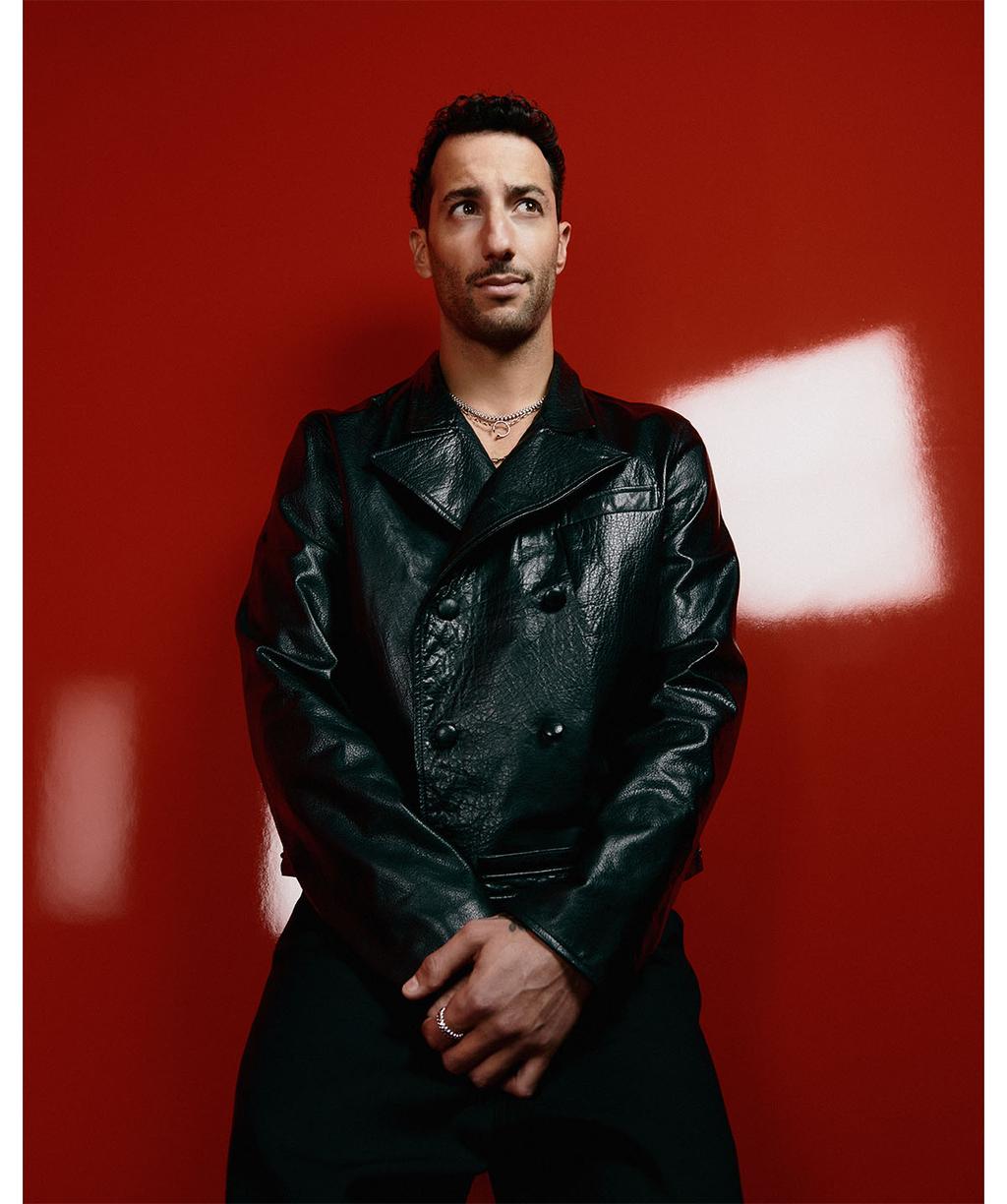 An adult male poses indoors against a red backdrop, wearing a black double-breasted jacket and jewelry, looking off-camera with a neutral expression.