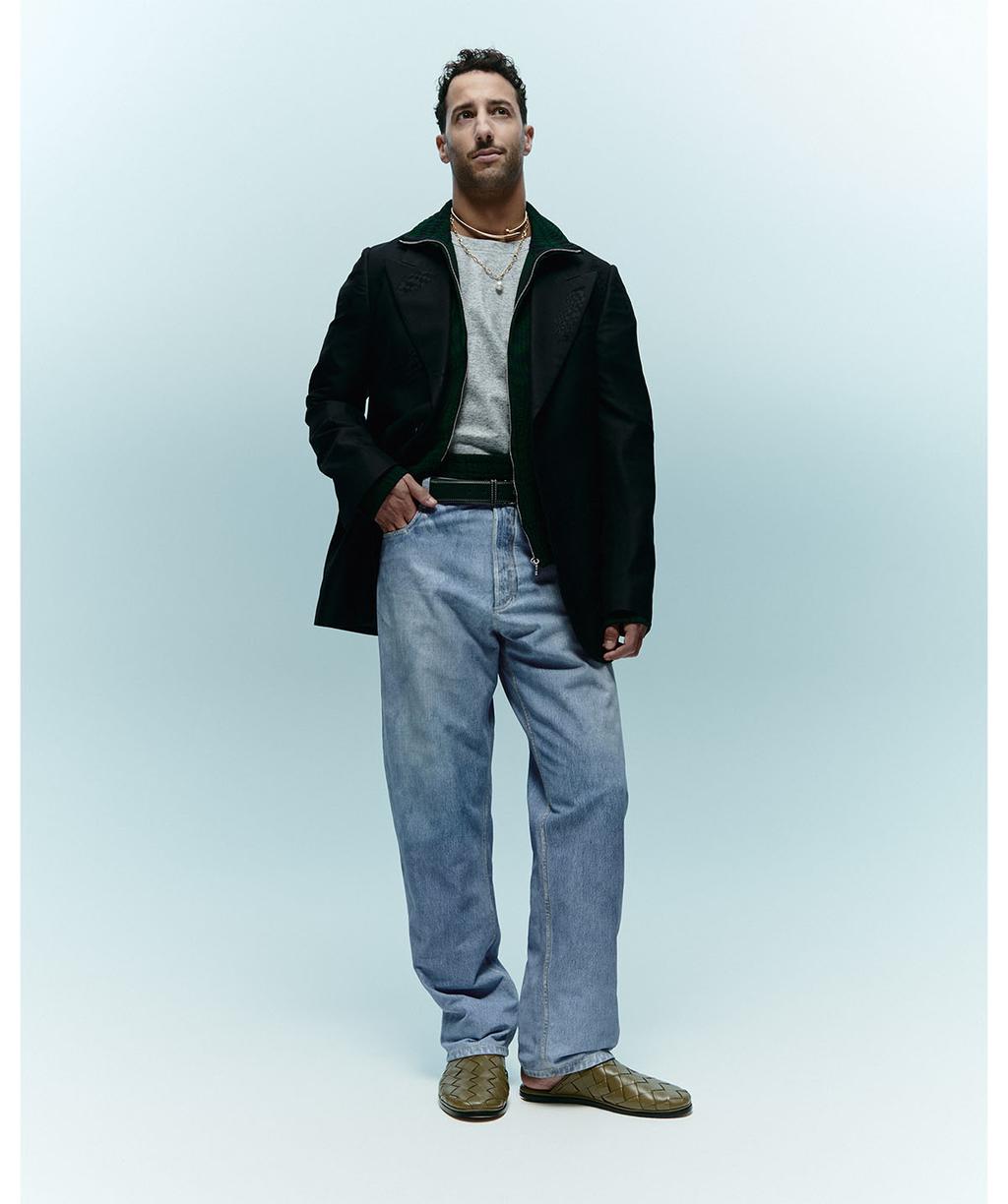 Full-length portrait of a smiling adult male wearing a blazer, jeans, and casual footwear, standing in a photo studio.
