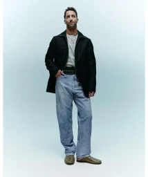 Full-length portrait of Daniel Ricciardo wearing a dark jacket over a grey t-shirt and light blue jeans against a light gradient studio backdrop