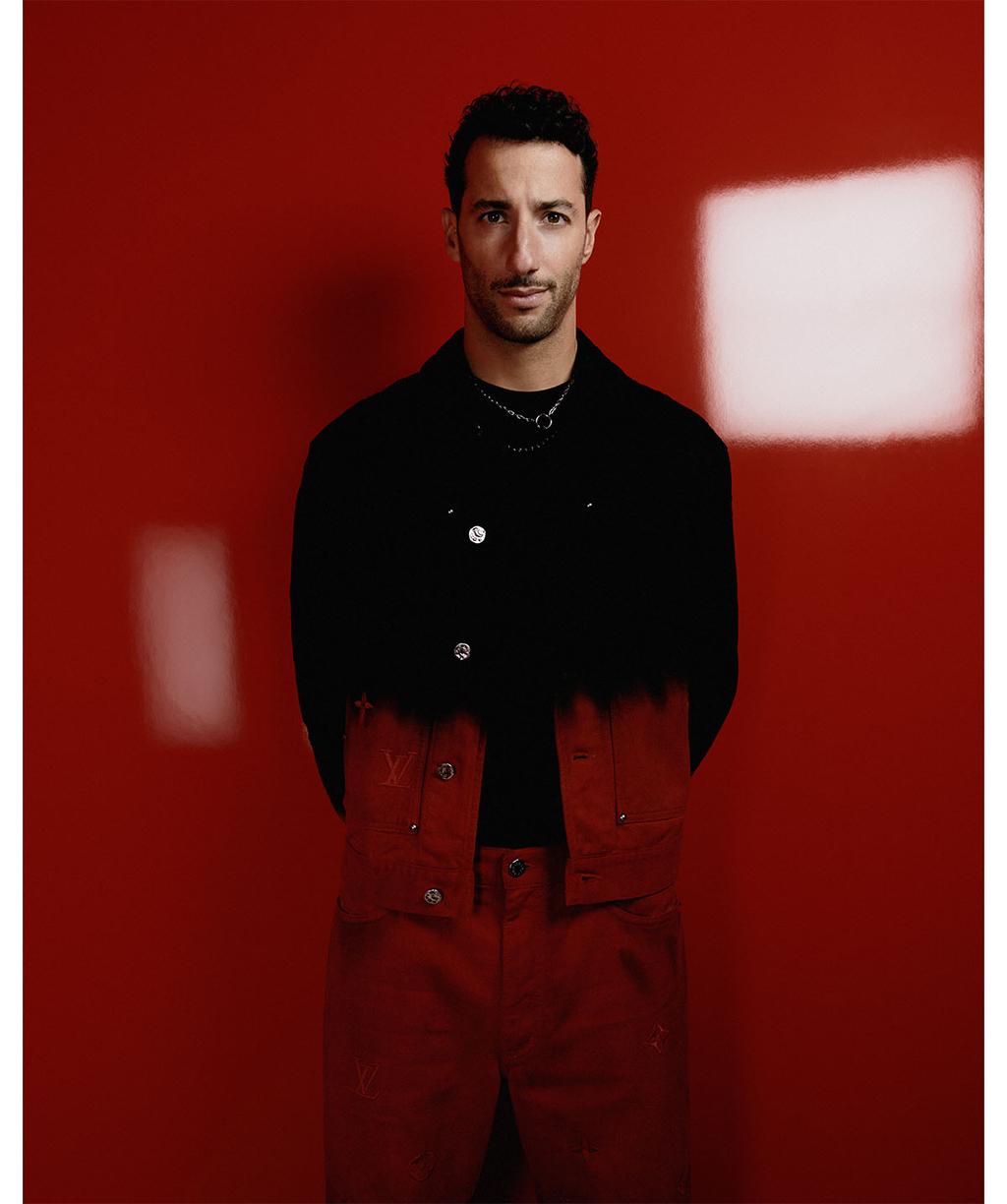 Studio portrait of an adult male wearing a black jacket with dark red pants standing against a red background with soft window lighting.