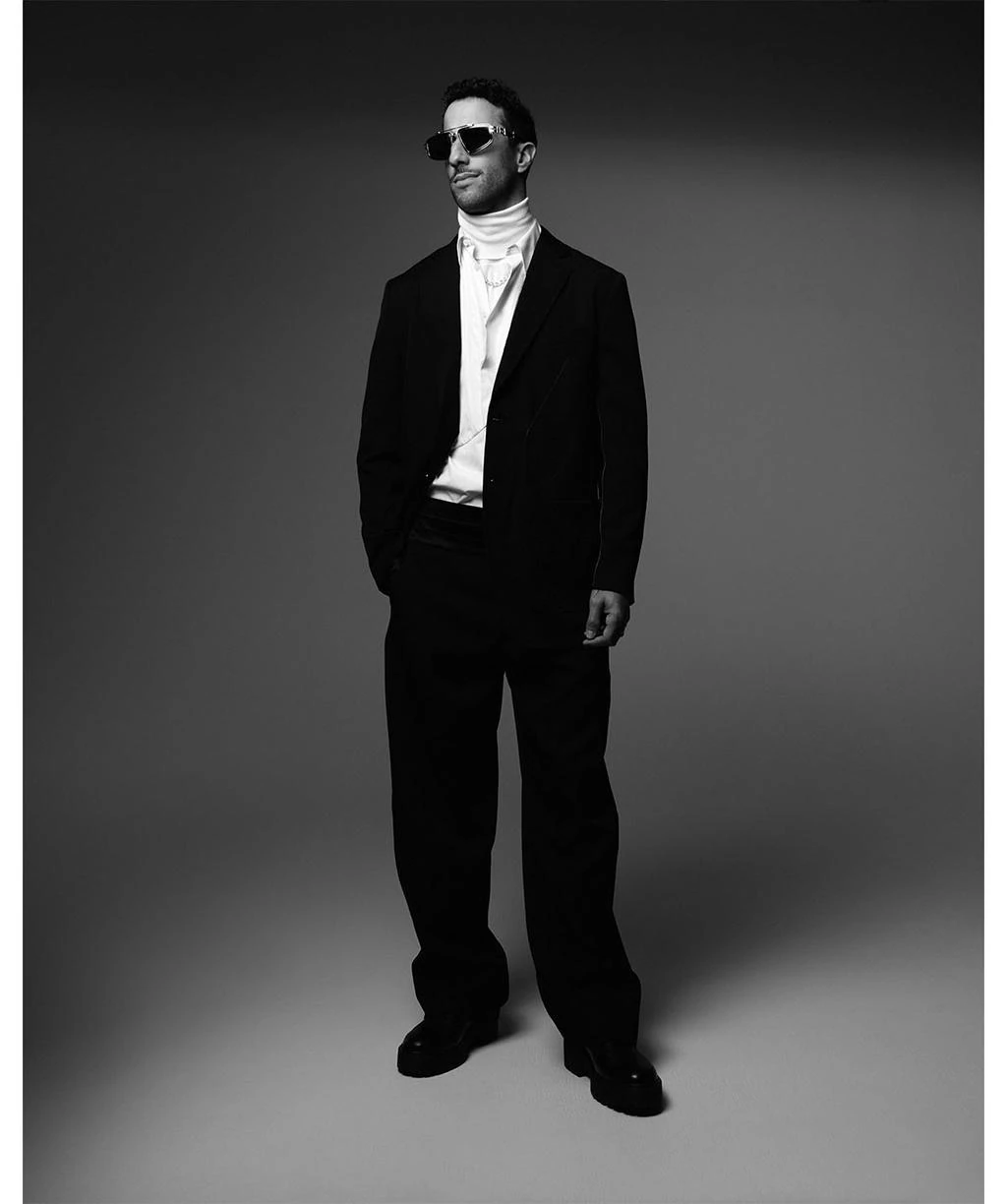 Monochrome full-length portrait of a man wearing a dark suit, sunglasses, and layered white shirt against a neutral studio backdrop.