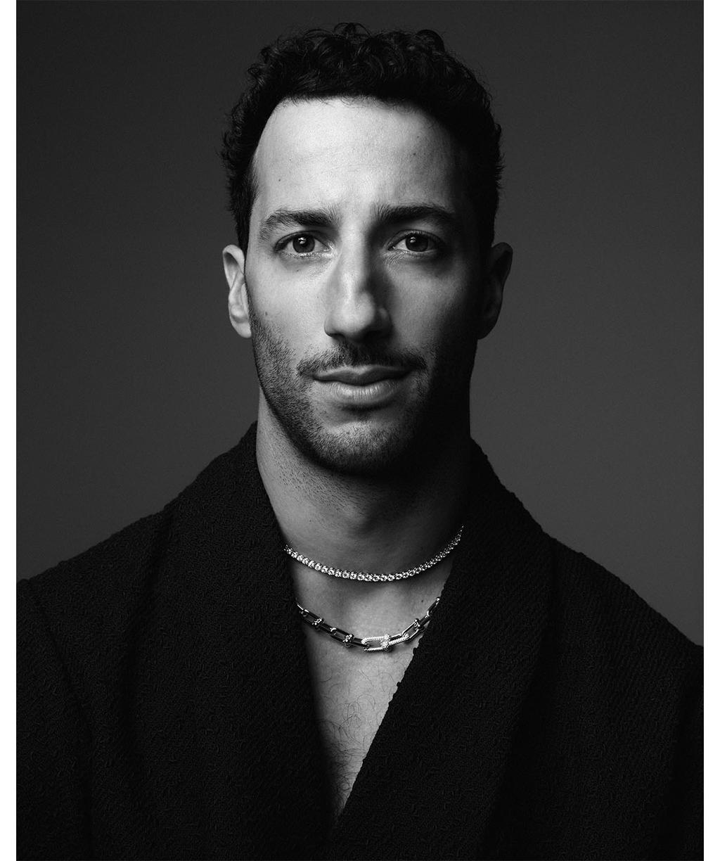 Monochromatic portrait of adult male with short dark hair, silver necklace, dark jacket against dark background, looking at camera.