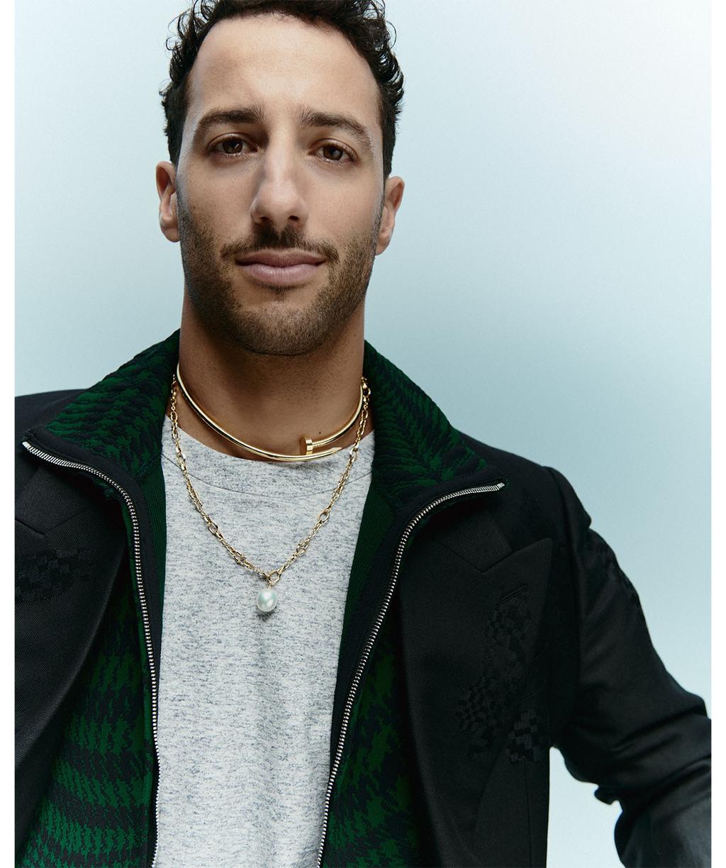 Close-up portrait of Daniel Ricciardo, wearing a t-shirt, jacket, and jewelry against a light blue studio backdrop.