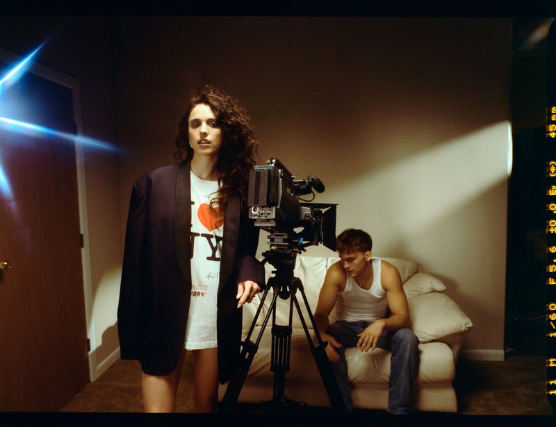 Woman in 'I love NY' t-shirt and blazer poses next to vintage camera on tripod in a dimly lit interior, man seated in background.