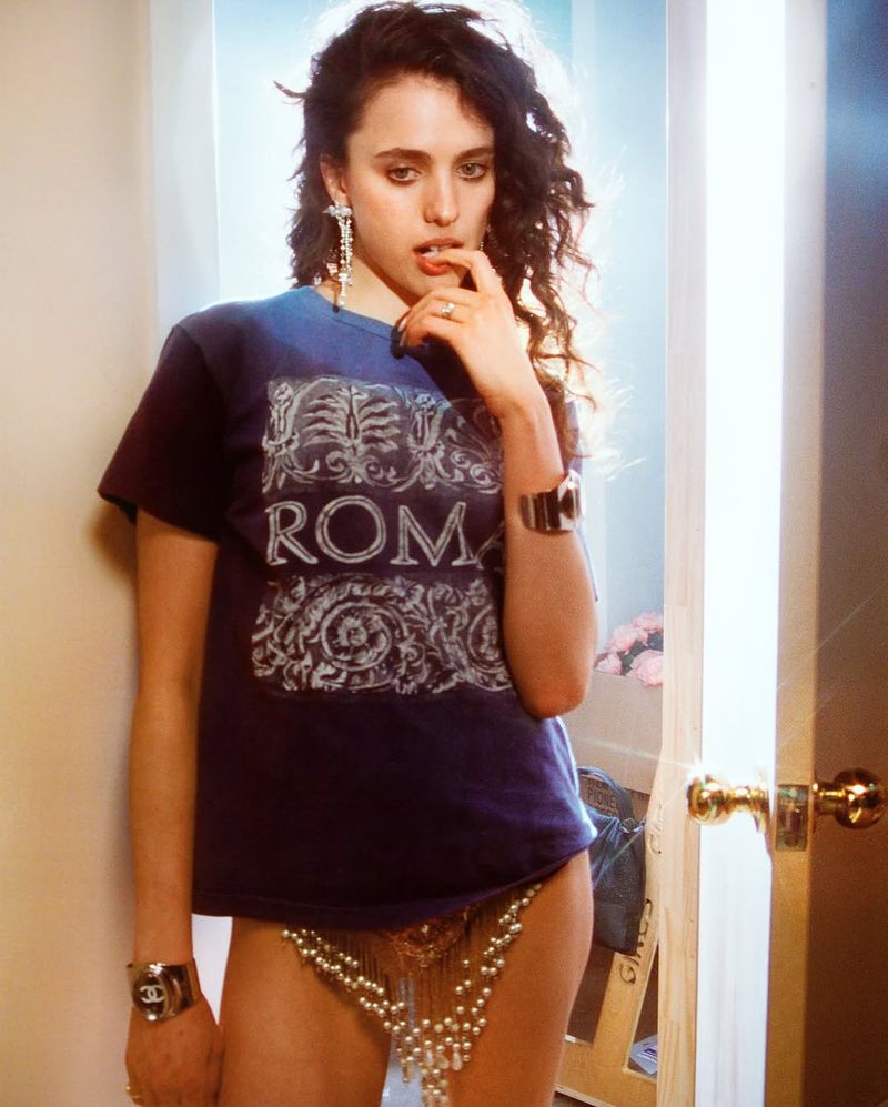 Young adult woman with curly hair poses indoors wearing a blue T-shirt with "Roma" graphic, large earrings and cuff bracelets.