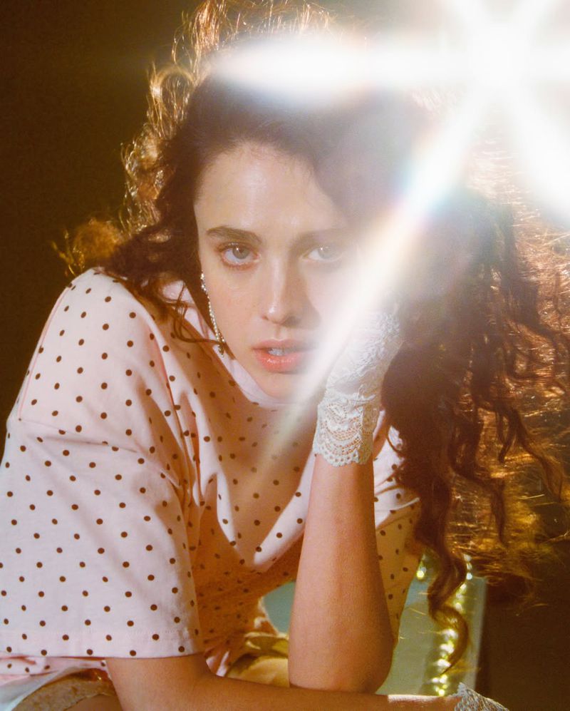 Portrait of a woman with wavy brown hair wearing a pink polka dot shirt with white lace gloves in front of a dark background.