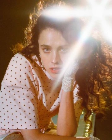 Portrait of a woman with wavy brown hair wearing a pink polka dot shirt with white lace gloves in front of a dark background.