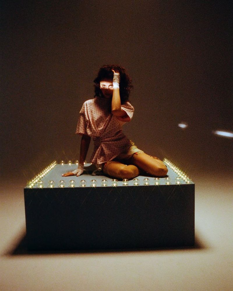 Margaret Qualley kneeling on an illuminated platform wearing a patterned top in an editorial portrait by Petra Collins for i-D Magazine