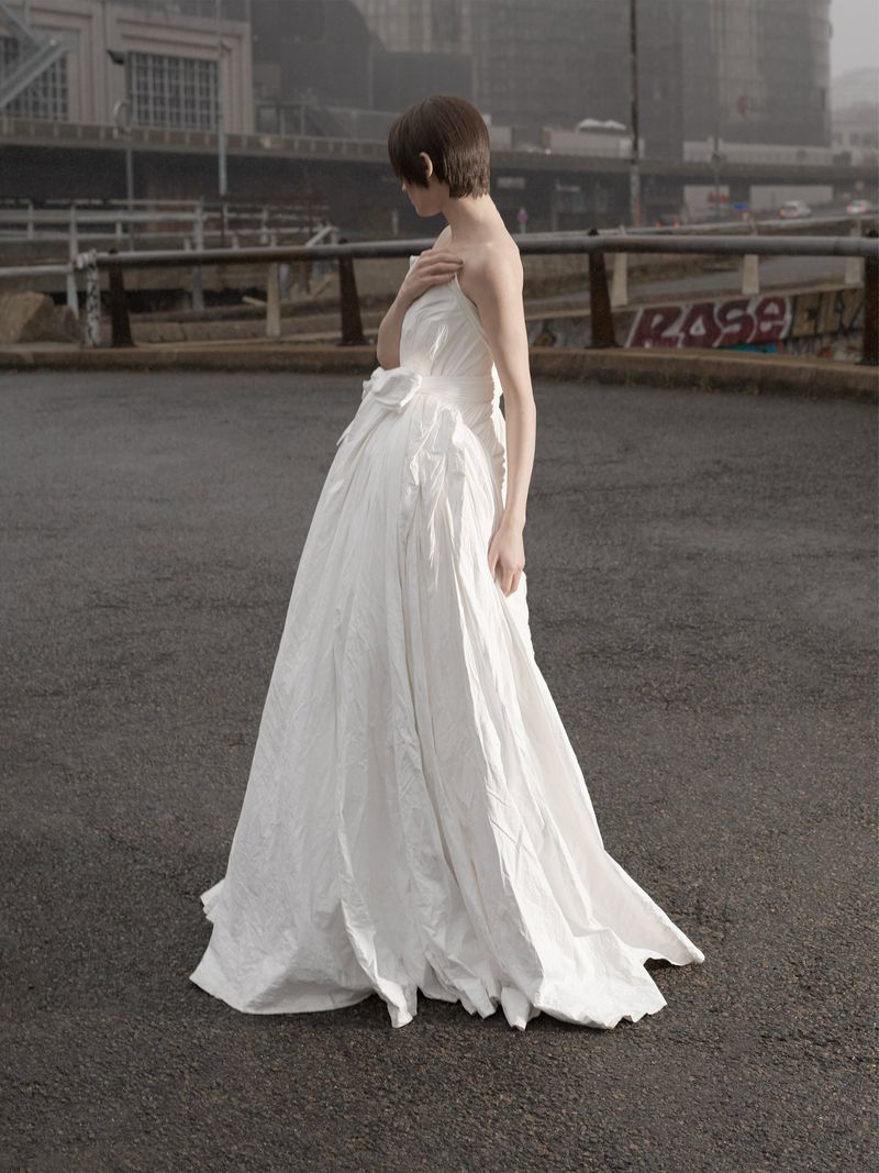 A woman in a white dress stands on an asphalt surface in an urban setting. Buildings and a bridge are visible in the background.