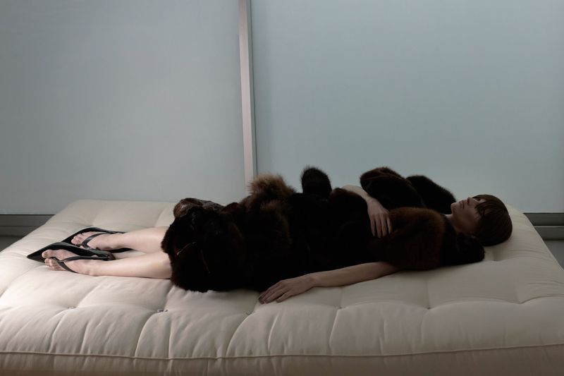 Model lying horizontally on a tufted white daybed wearing a dark fur jacket against a pale wall.