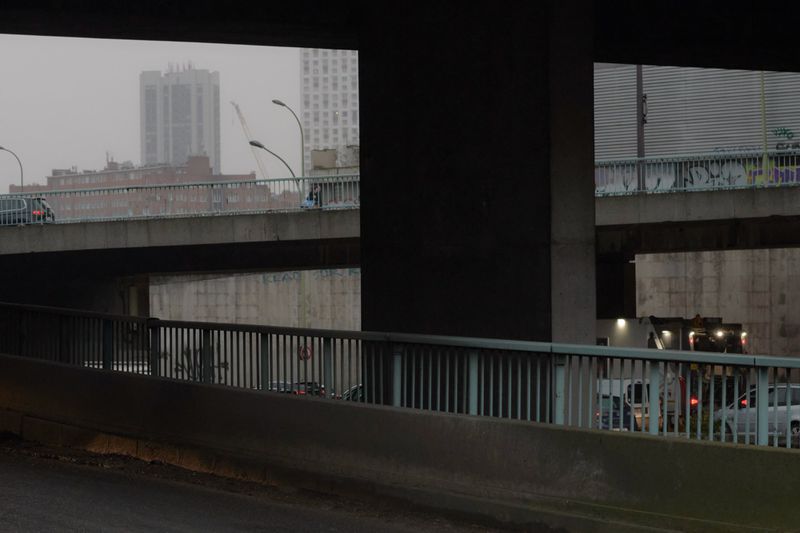 Cityscape featuring a concrete overpass with vehicle traffic, viewed from a low angle under an underpass, in an urban setting with overcast lighting.