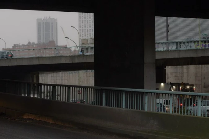 Cityscape featuring a concrete overpass with vehicle traffic, viewed from a low angle under an underpass, in an urban setting with overcast lighting.
