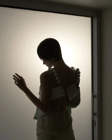 Back view of a slender adult woman with bobbed hair wearing a pearl strap top and loose-fitting pants in a studio environment.