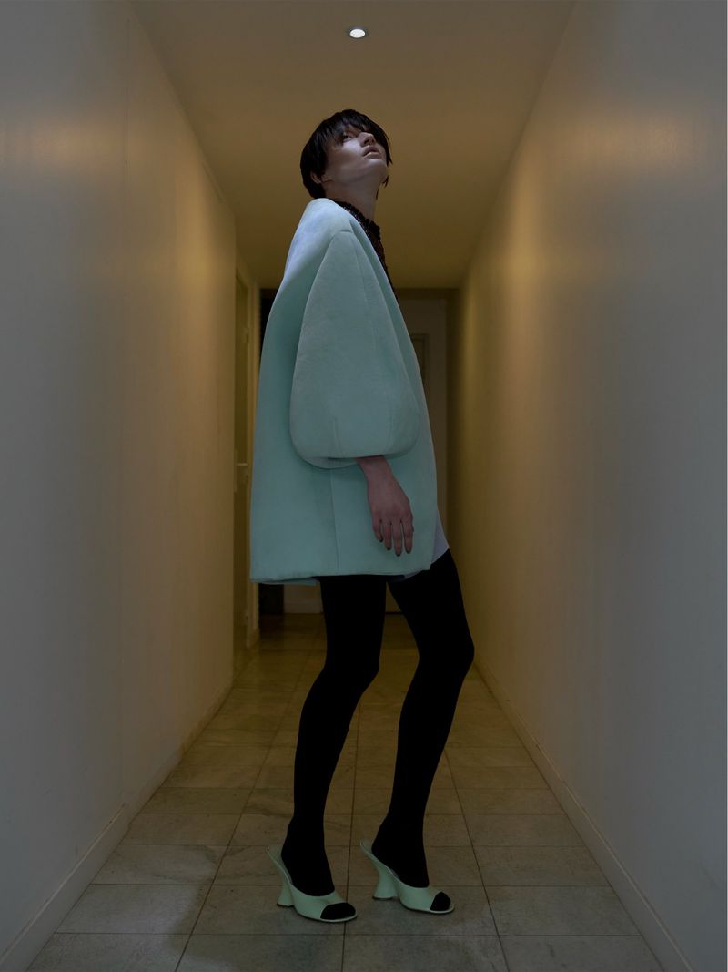 Adult female in a light green coat with black tights and heels stands in a hallway with yellow-tinted walls and a bright ceiling light.