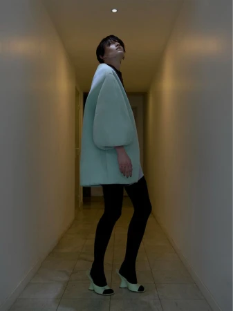 Adult female in a light green coat with black tights and heels stands in a hallway with yellow-tinted walls and a bright ceiling light.