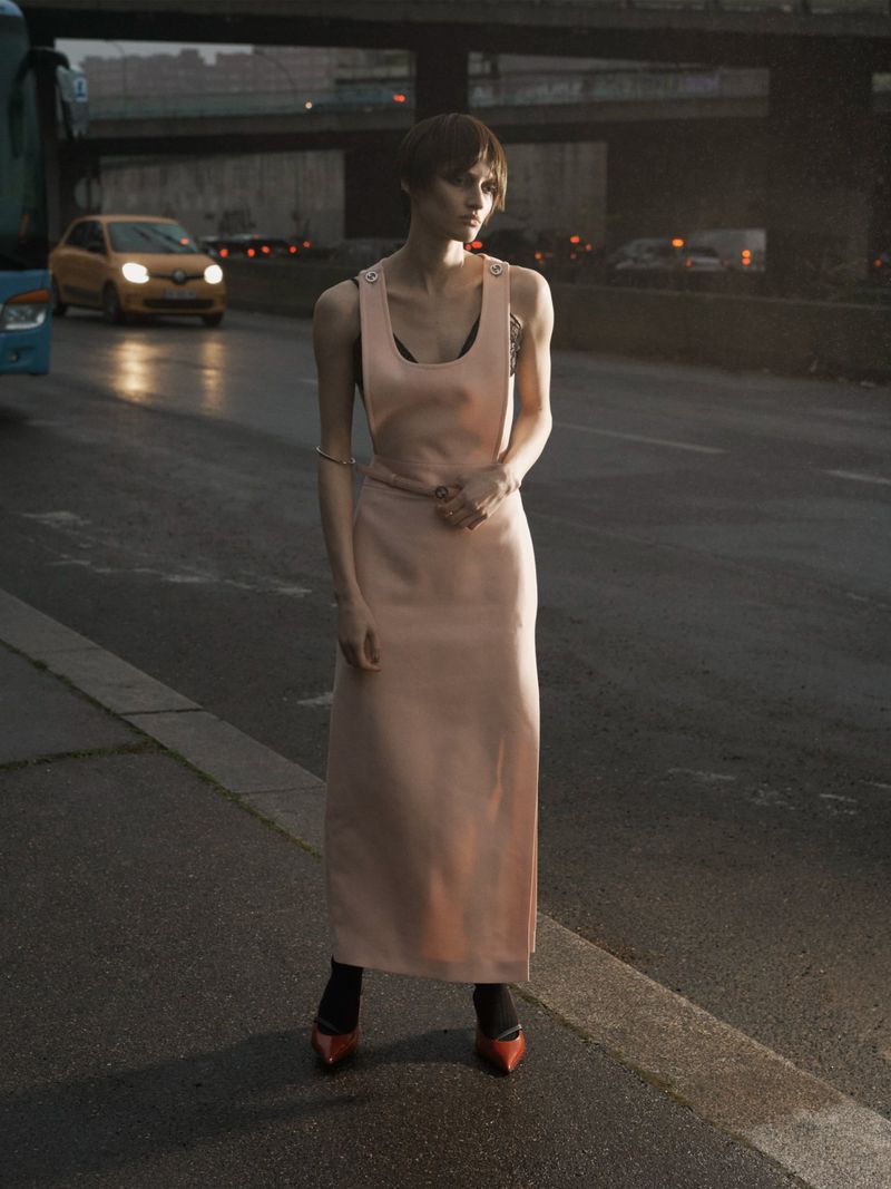 A female model in a pink dress stands on a city street with blurred traffic in the background, showcasing an urban fashion editorial.