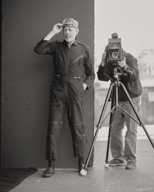 A photographer captures an image of a man in a jumpsuit and glasses with a large format camera indoors against a gray background.