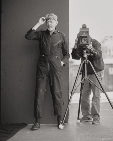 A photographer captures an image of a man in a jumpsuit and glasses with a large format camera indoors against a gray background.