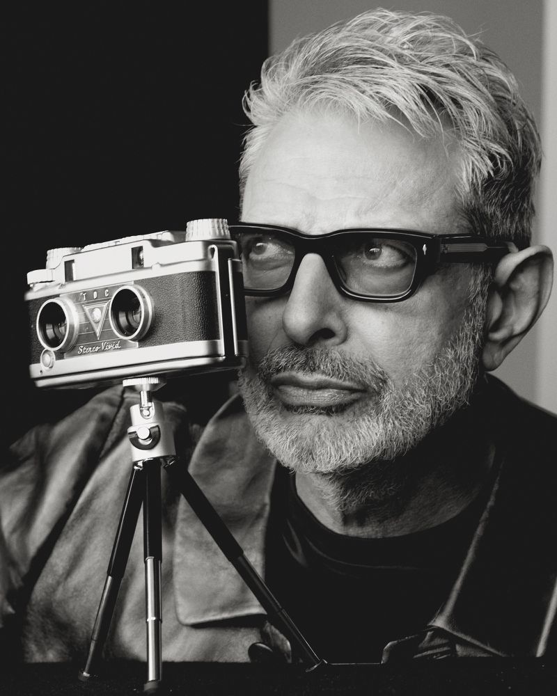 Monochrome portrait of a stylish man with glasses holding a vintage stereo camera on a tripod, set against a solid dark backdrop.
