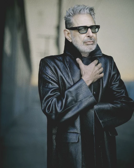 Portrait of Jeff Goldblum wearing sunglasses and a black leather coat, standing in front of a gray concrete wall.