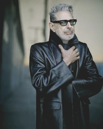 Portrait of Jeff Goldblum wearing sunglasses and a black leather coat, standing in front of a gray concrete wall.