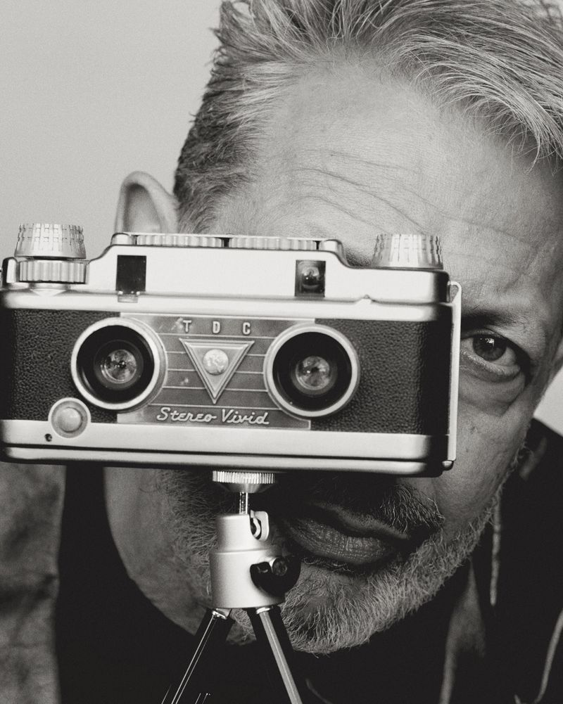 Black and white portrait of a man holding a vintage twin-lens stereo camera on a tripod, with his eyes looking directly at the camera.