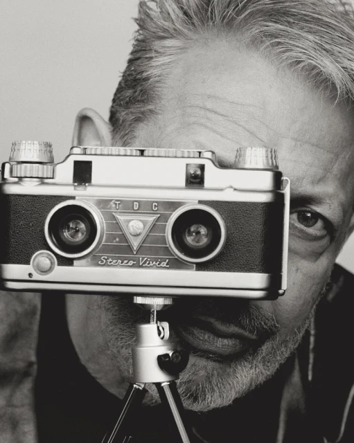 Black and white portrait of a man holding a vintage twin-lens stereo camera on a tripod, with his eyes looking directly at the camera.