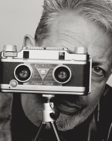 Black and white portrait of a man holding a vintage twin-lens stereo camera on a tripod, with his eyes looking directly at the camera.