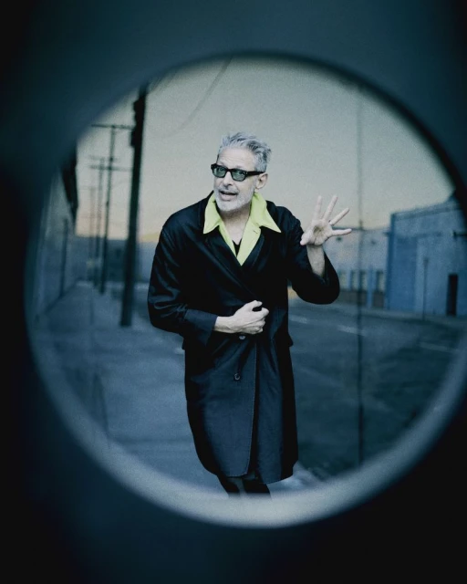 Man with grey hair in black coat and sunglasses stands on street, captured through circular frame, with urban background and subdued lighting.
