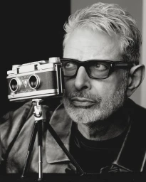 Jeff Goldblum wearing thick black glasses and looking past a vintage stereo camera mounted on a mini tripod in a monochrome portrait.