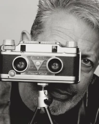 Jeff Goldblum holding a vintage TDC Stereo Vivid camera up to his face against a plain background in a black and white portrait