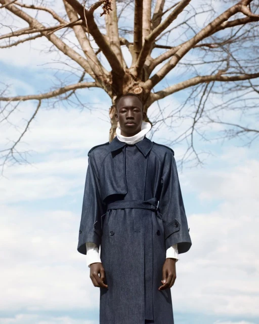 Portrait of a man in a trench coat standing beneath a bare tree against a sky with clouds. Fashion shoot with a serious expression.