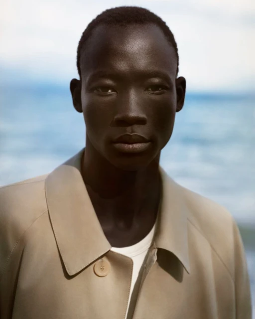 Headshot of a man with dark skin wearing a beige trench coat against a blurred background of a light blue ocean and sky.