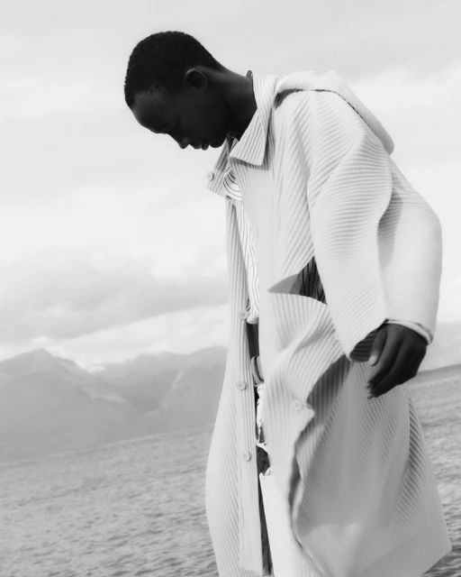 Black and white portrait of an African male model in a hooded jacket on a beach with mountains in the background on a cloudy day.