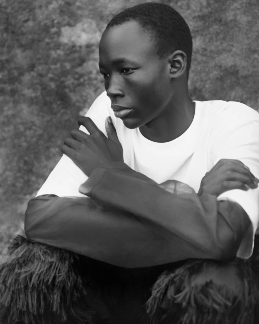 Monochrome portrait of a black man in a white shirt, posed with arms crossed in front of a blurry dark grey mottled background.