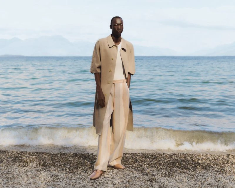Man poses on a pebble beach in a beige coat and striped pants against a water and mountain background on a sunny day.