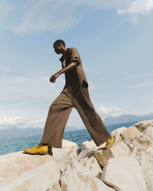 An adult male strides across rocks near the sea in a brown striped suit and yellow shoes against a blue sky with light clouds.