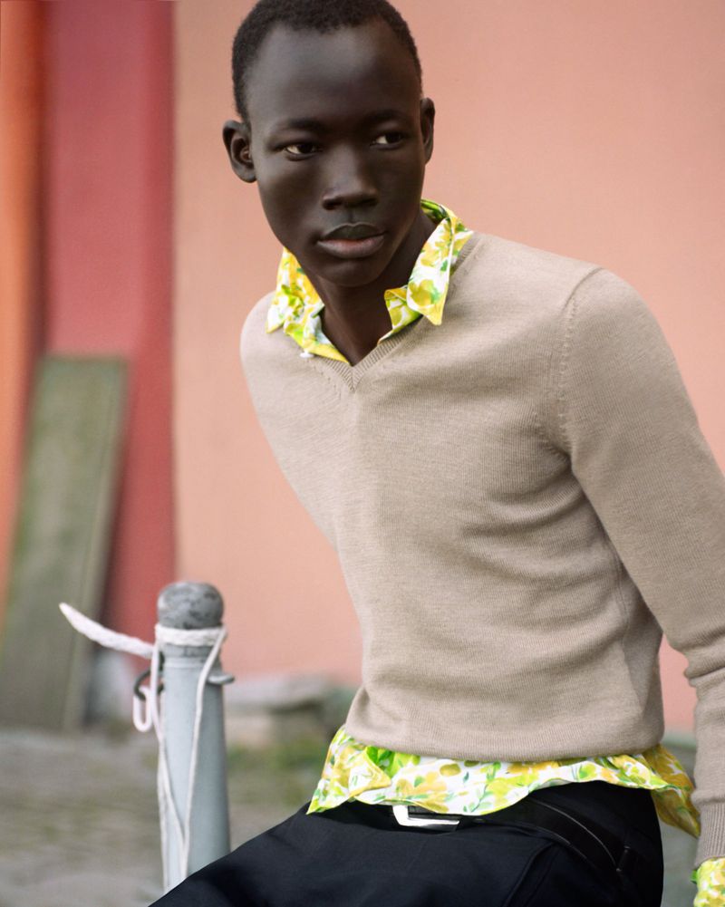 Portrait of a dark-skinned adult male wearing a tan sweater and a colorful button-down shirt, leaning against a gray metal pole outdoors.