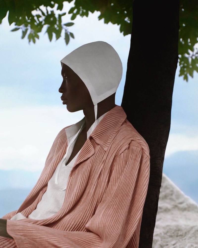 Fashionable woman in pink jacket and white hat poses next to tree with sky and pyramid structure in the background on bright day.