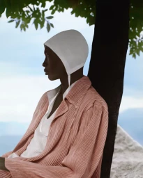 Model in pink pleated jacket and white bonnet leaning against a tree trunk outdoors.