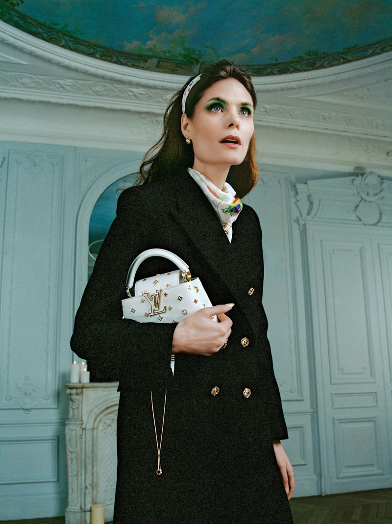 A woman in a black coat holds a patterned handbag in a bright room with elegant molding. She wears a headband and bright eyeshadow.