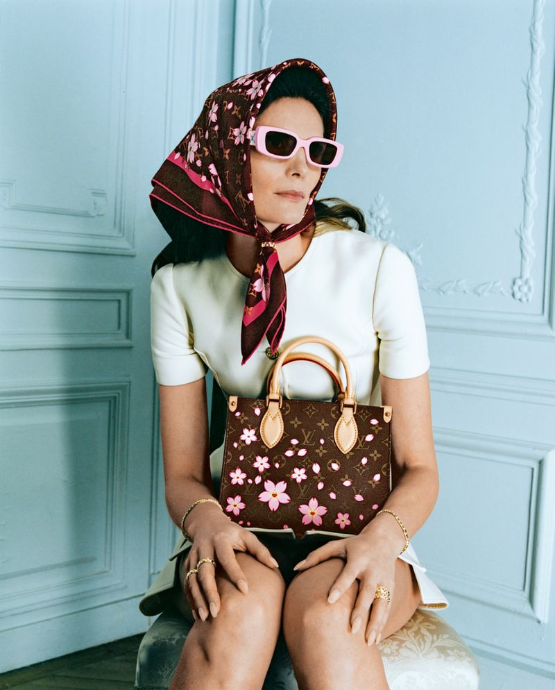 Elegant woman in white dress, sunglasses, and floral headscarf sits with a patterned handbag in a brightly lit indoor setting.